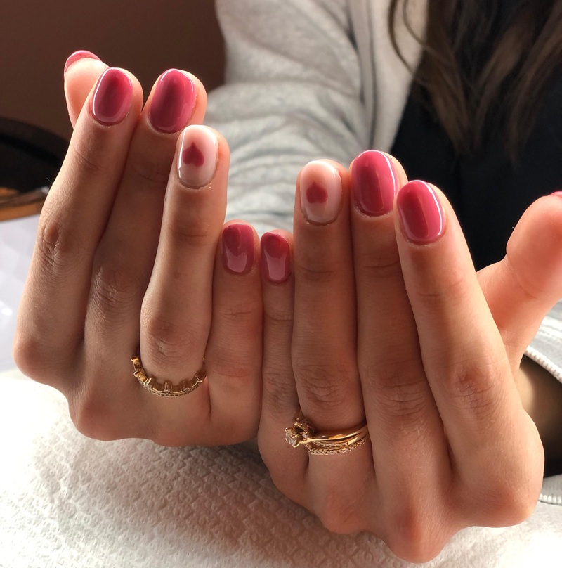 Mani con unghie corte smaltate rosa lampone, anulari nude con cuoricino rosso e anelli dorati su asciugamano bianco